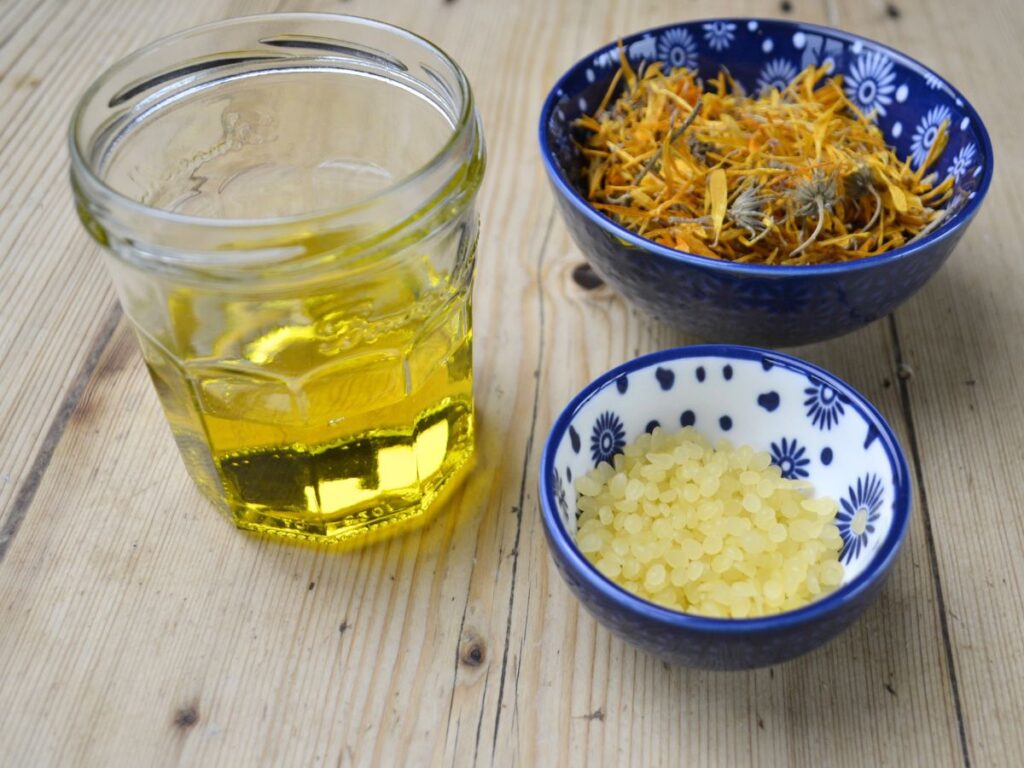 A jar with oil a bowl with dryvcalendula flowers and a bowl with beeswax pellets