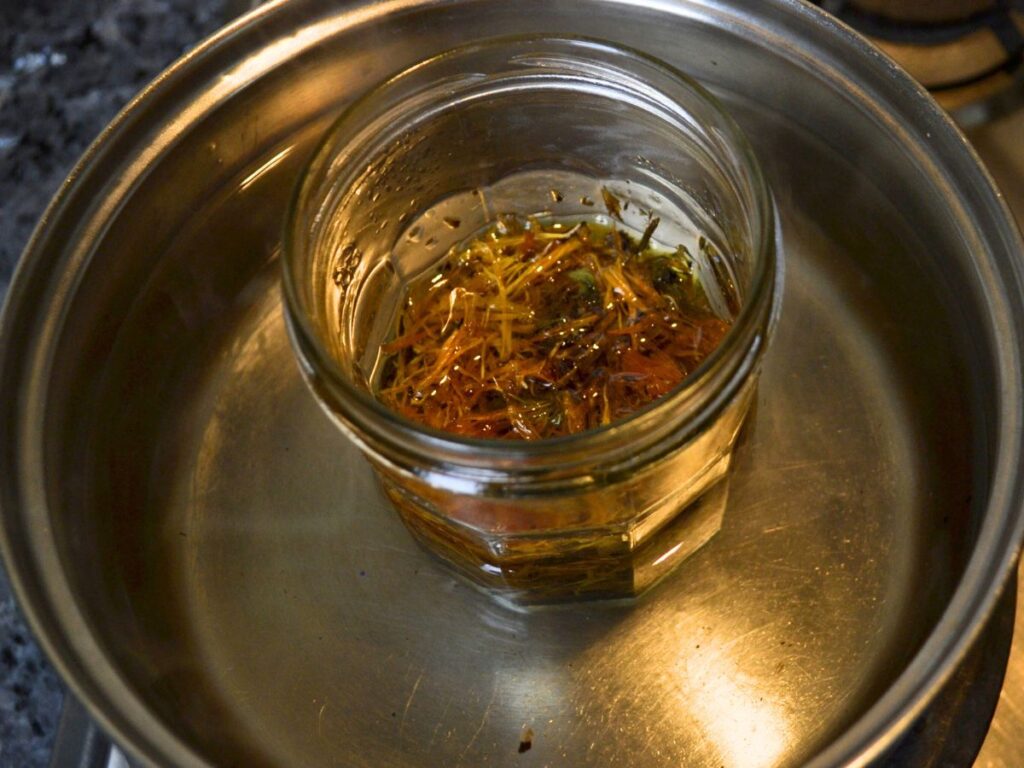 A glass jar with oil and dry calendula flowers on a soucepan with water