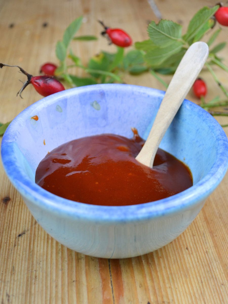 A small blue bowl with Rosehip jam, a wooden spoon, and a wild rose branch in the background.