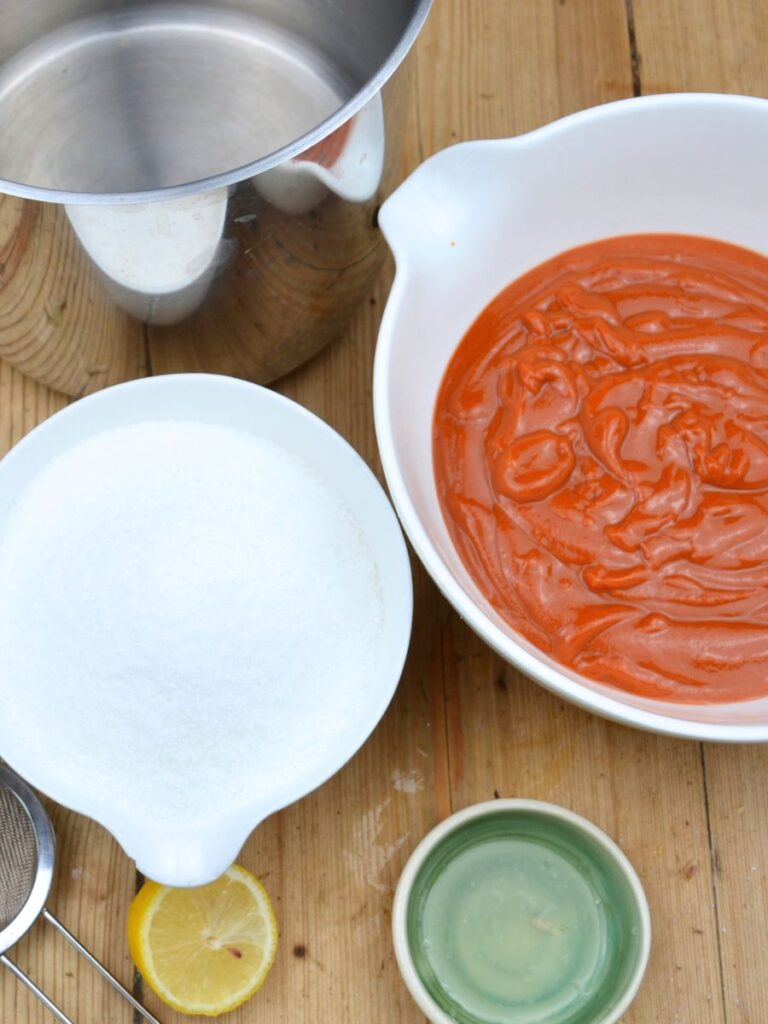 A pot, a bowl of rosehip puree, a bowl of sugar, half a lemon, and a small plate.