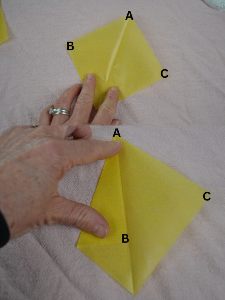 A hand holding a square of kite paper with a center crease and folding one point to the crease.